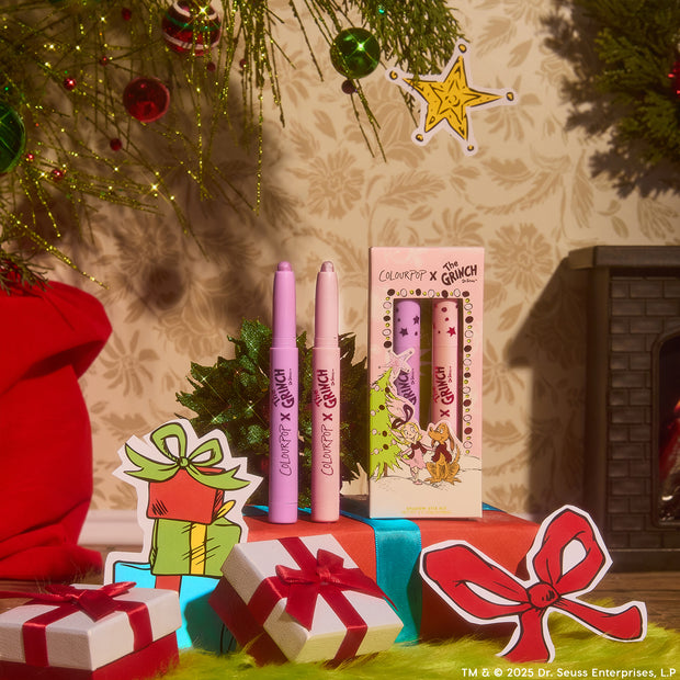 ColourPop Liked Christmas a Lot… Shadow Stix Set displayed upright - two lilac eyeshadow sticks beside a boxed trio in pastel packaging - surrounded by ornaments, wrapped gifts, and a fireplace in a cheerful Grinch-themed holiday scene.