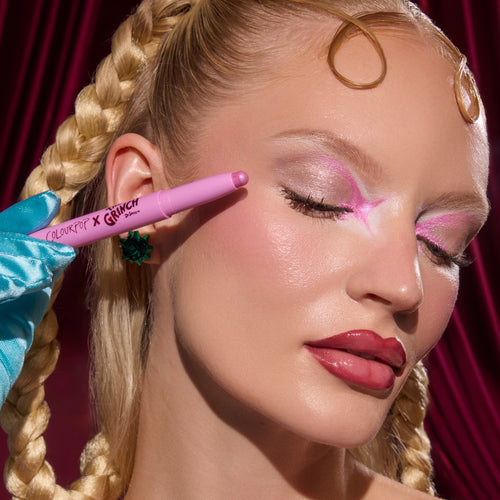 ColourPop x Grinch Shadow Stix in shimmery pink is held by a teal glove to a model’s eyelid, demonstrating creamy eyeshadow application over pink-and-silver glam makeup, with braided hair, green bow earring, and a red velvet backdrop.