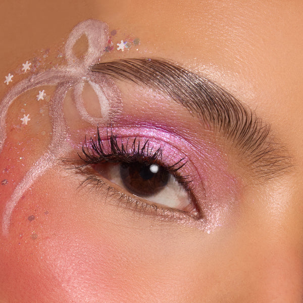 ColourPop Who-Hash Glitter Gel sparkling across the eyelid and brow bone, layering pink iridescent shimmer with star confetti and white ribbon swirls for a festive eye look in a close-up beauty shot.