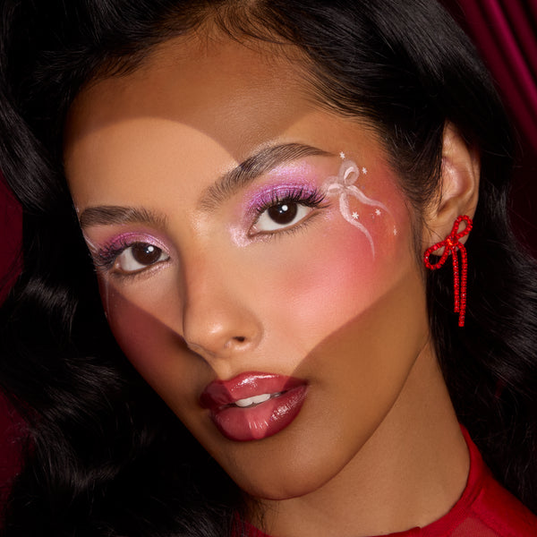 Glossy berry-red lips showcase the ColourPop Pouty Grinch Lip Set, worn by a model with pink shimmer eyeshadow, white ribbon face art, and a red bow earring against a deep red backdrop.
