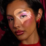Model wears shimmering pink lids and icy inner-corner highlight created with the ColourPop Liked Christmas a Lot… Shadow Stix Set, accented by blush, glossy red lips, a heart-shaped light, and a red bow earring against a deep red backdrop.