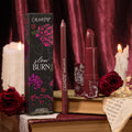 ColourPop Lux Lipstick in Slow Burn, a deep rose‑burgundy bullet with matching lippie pencil, stands beside a floral box on stacked books, framed by red roses and melting candles against a rich burgundy velvet backdrop.