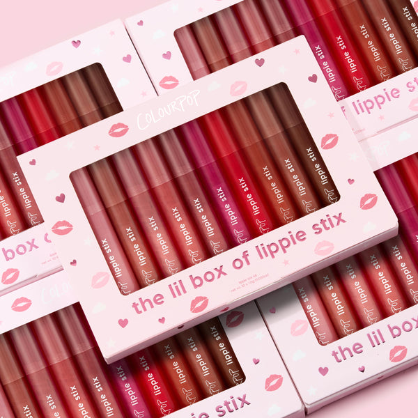 The Lil Box of Lippie Stix ColourPop Lippie Stix set displayed in windowed pink boxes, showcasing assorted nude, pink, and red shades; multiple kits stacked on a pastel pink background with hearts and kiss prints.