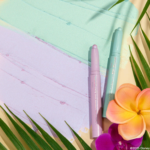 Two pastel ColourPop Shadow Stix, Cute and Feisty, in mint and lilac, rest on matching shimmery swatches, framed by tropical palm leaves and pink-orange plumeria and purple orchid, showcasing the creamy twist-up eyeshadow duo.