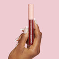 ColourPop LAX Ultra Matte Lip liquid lipstick held upright by a hand with white‑tipped nails, showing a deep burgundy shade in a clear tube with a pink star-print cap against a solid pastel pink background.