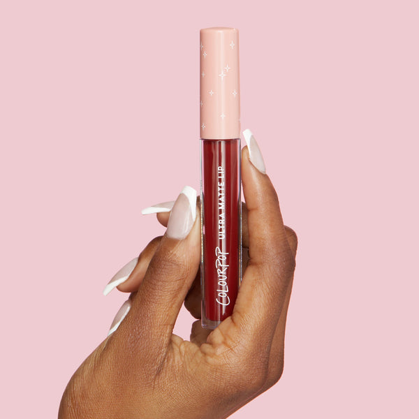 ColourPop LAX Ultra Matte Lip liquid lipstick held upright by a hand with white‑tipped nails, showing a deep burgundy shade in a clear tube with a pink star-print cap against a solid pastel pink background.
