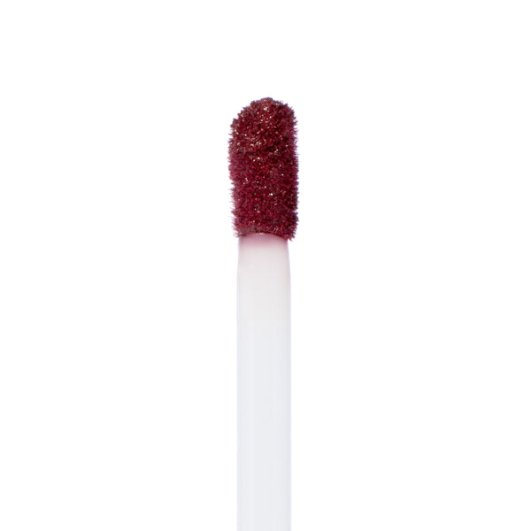 Doe-foot applicator coated with ColourPop LAX Ultra Matte Lip, a deep burgundy liquid lipstick, depositing pigment in a clean macro close-up against a white studio background.