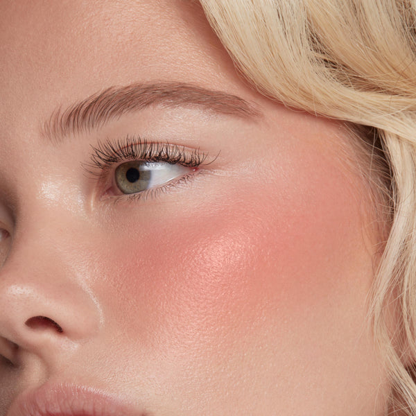 ColourPop Liquid Blush in Super Shy sweeping a dewy peach‑pink flush across the cheekbone, blended to a soft luminous sheen; extreme close-up shows natural lashes and brow with blonde strands framing the face.