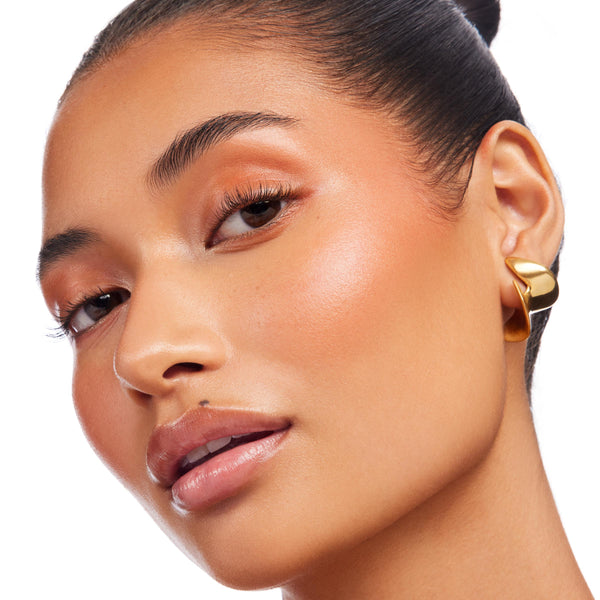 Model wears ColourPop Lite Stix Sunlight, swiped on cheekbones for a dewy peach-gold glow, paired with warm copper eyeshadow, glossy nude lips, and a sculptural gold earring in a clean studio close-up.