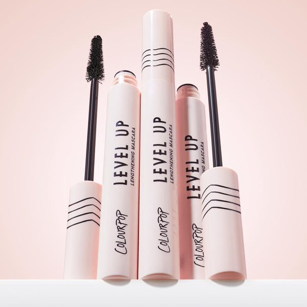 ColourPop Lengthening Mascara Level Up in Black stands upright with two uncapped wands revealing dense bristles, pale pink tubes marked with black stripes and logo on a soft pink backdrop, showcasing the sleek lash-lengthening product.