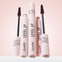 ColourPop Level Up Lengthening Mascara in Brown stands upright, with two open tubes showing slim bristle wands; pale pink packaging with black lines and logos grouped on a white surface against a soft pink backdrop.
