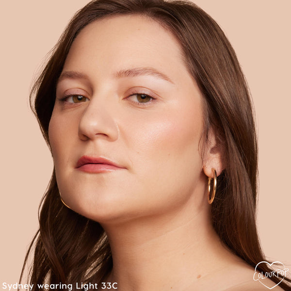 ColourPop Pretty Fresh Creamy Concealer, shade Light 33 C, evening complexion and brightening under eyes on a model; close-up shows a natural satin finish with a neutral-cool tone, set against a peach backdrop with gold hoop earrings.