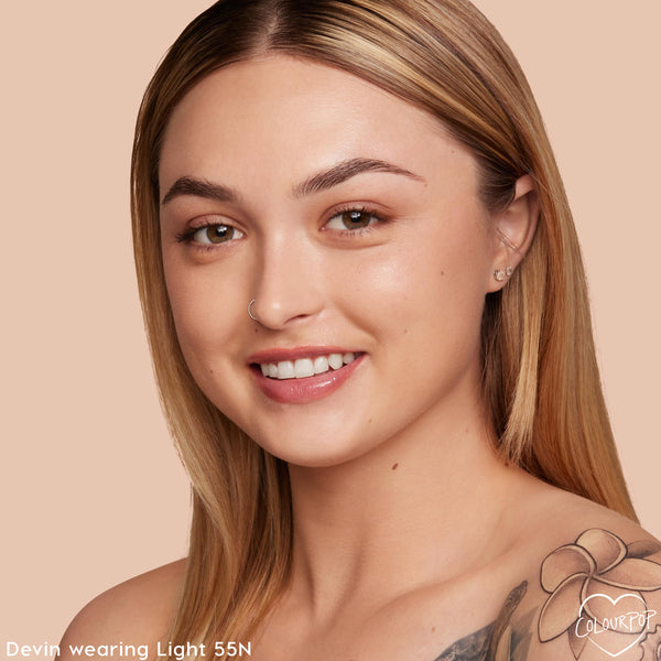 Model wearing Light 55 N neutral shade, showing smooth, dewy medium coverage from ColourPop Hyaluronic Hydrating Foundation in a close-up smile against a peach studio background.