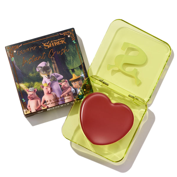 Fairytale Things ColourPop Lip and Cheek Balm sits open in a translucent neon-yellow square compact with an embossed S, revealing a red heart-shaped pan; beside it, the Shrek collab carton shows fairytale characters on a white studio background.