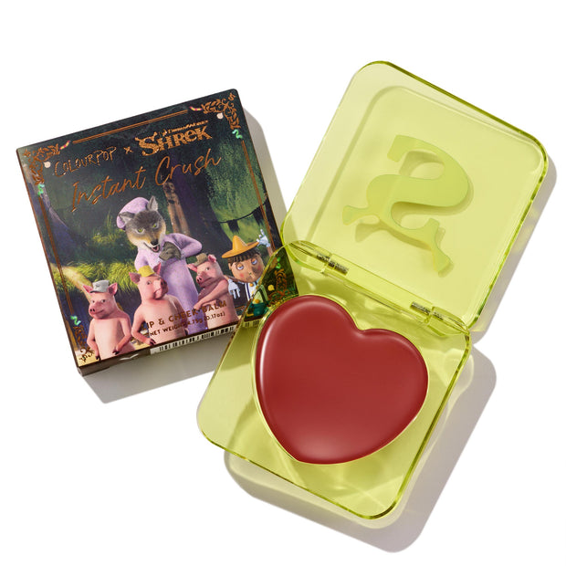 Fairytale Things ColourPop Lip and Cheek Balm sits open in a translucent neon-yellow square compact with an embossed S, revealing a red heart-shaped pan; beside it, the Shrek collab carton shows fairytale characters on a white studio background.