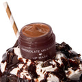 Chocolate Milkshake ColourPop Lip Mask jar sits open, revealing a glossy caramel-brown formula, resting atop whipped cream drizzled with chocolate and dotted with chunks, with a striped straw, showcasing a dessert-inspired hydrating lip treatment.
