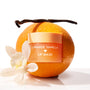Orange Vanilla ColourPop Lip Mask jar gleaming with a peach-gold lid, sitting against a fresh orange as a vanilla bean and white blossom frame it, styled on a bright white background to highlight the citrus-vanilla scent and hydrating treatment.
