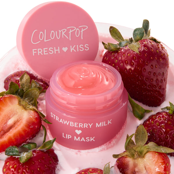 Open jar of ColourPop Lip Mask, Strawberry Milk, showing a glossy pink swirl, resting in a bowl of strawberry milk with fresh strawberries; matching pink lid reading "ColourPop Fresh Kiss" set beside it.