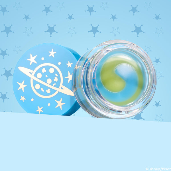 Clear jar of The Chosen One ColourPop Lip Mask showing a yin-yang swirl of green and blue balm, resting open beside a sky-blue lid printed with a ringed planet and stars, set on a light blue background with star-badge patterns.