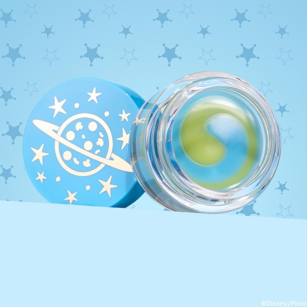 Clear jar of The Chosen One ColourPop Lip Mask showing a yin-yang swirl of green and blue balm, resting open beside a sky-blue lid printed with a ringed planet and stars, set on a light blue background with star-badge patterns.
