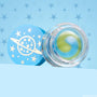 Clear jar of The Chosen One ColourPop Lip Mask showing a yin-yang swirl of green and blue balm, resting open beside a sky-blue lid printed with a ringed planet and stars, set on a light blue background with star-badge patterns.