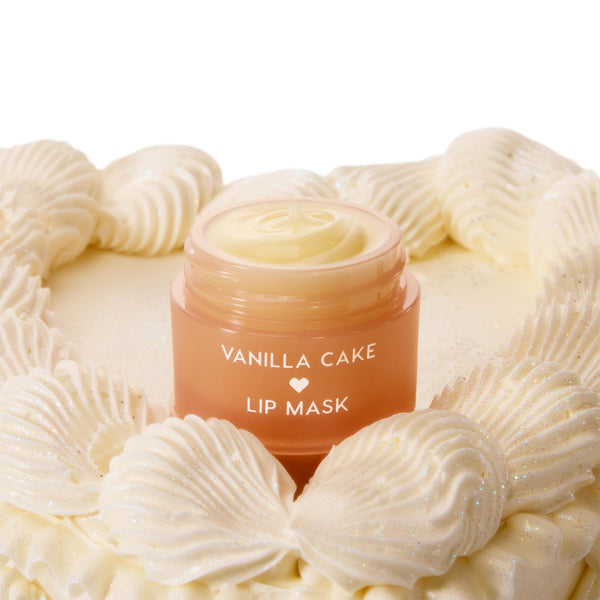 Vanilla Cake ColourPop Lip Mask jar, lid off to reveal creamy pale-yellow balm, set atop a vanilla cake with glittery piped frosting; peach ombre container with white lettering and a heart icon.
