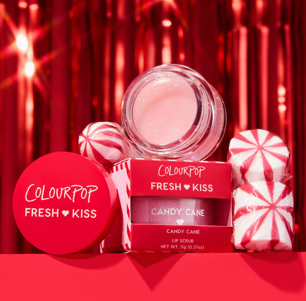 ColourPop Fresh Kiss Candy Cane lip scrub jar and red box showcasing a shimmery pink formula, staged with peppermint candies against a festive red tinsel backdrop; holiday lip exfoliator, 11 g (0.37 oz).
