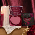 Heart-shaped Love Interest compact open to reveal rosy mauve balm, displayed with floral “Instant Crush” box, lit candles, a torn book page, and dried roses on a wood table - ColourPop Lip and Cheek Balm for lips and cheeks.