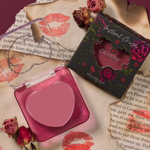 Love Interest ColourPop Lip and Cheek Balm shows a heart-shaped rosy mauve pan in a translucent pink compact, opened beside its black floral box with heart cutout, styled on torn book pages with red lipstick kiss prints and dried roses.