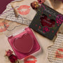 Ravished ColourPop Lip and Cheek Balm sits open, revealing a heart-shaped mauve tint in a translucent pink compact, beside its floral black box with heart window, styled on torn pages with lipstick kisses and dried roses.