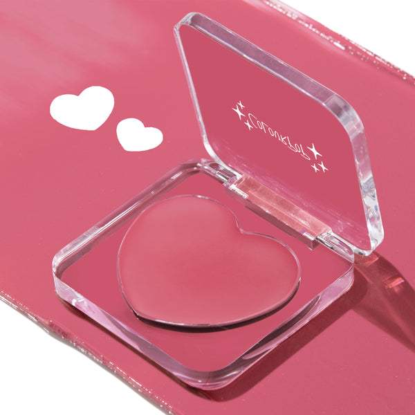 Heart-shaped pink cream in a clear acrylic compact sits open, showcasing the Verified ColourPop Lip and Cheek Balm on a glossy rose background with two white heart graphics, perfect for buildable blush or tinted lip color.