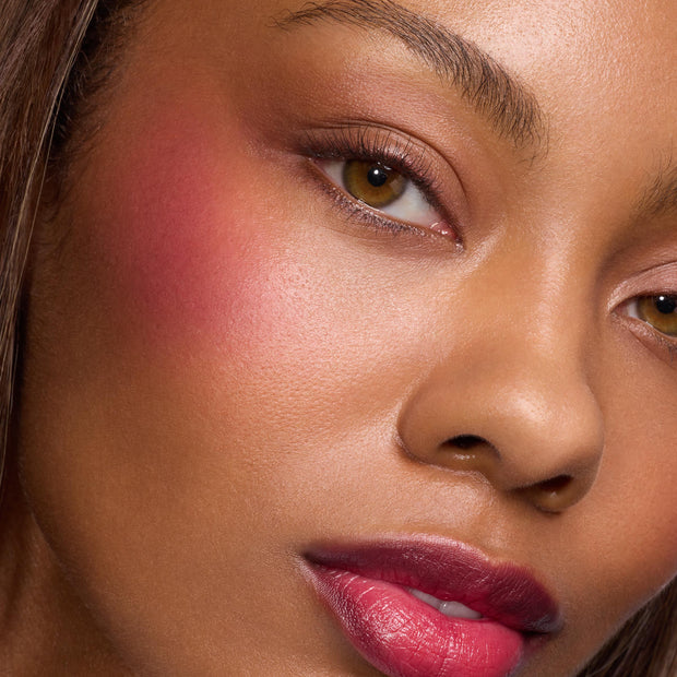Model wearing ColourPop Lip and Cheek Balm in Ravished, showcasing a rosy berry flush on the cheeks and glossy tinted lips, highlighting the buildable, dewy finish in a close-up beauty shot.