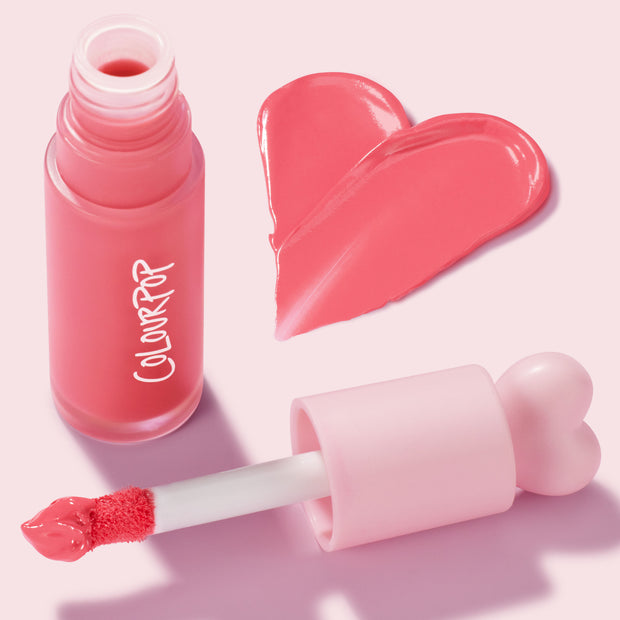 ColourPop Liquid Blush Angel Face shade displaying a glossy coral-pink heart swatch, with an open frosted bottle labeled COLOURPOP and a doe-foot applicator topped by a pink heart cap, set on a light pink backdrop.