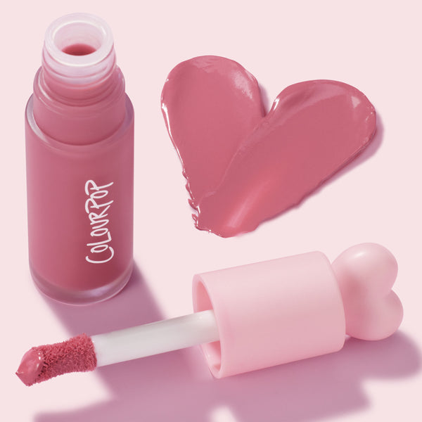 IKTR ColourPop Liquid Blush swatched in a rosy pink heart, while a frosted bottle with white ColourPop logo stands open and a doe-foot applicator with heart-shaped pink cap rests nearby, highlighting a creamy, dewy finish on a pastel pink backdrop.