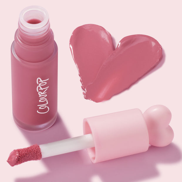 IKTR ColourPop Liquid Blush swatched in a rosy pink heart, while a frosted bottle with white ColourPop logo stands open and a doe-foot applicator with heart-shaped pink cap rests nearby, highlighting a creamy, dewy finish on a pastel pink backdrop.