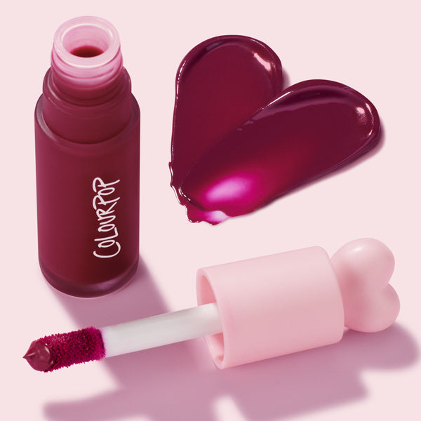 ColourPop Liquid Blush Royal Flush, an open frosted berry bottle with white logo and pink heart-top cap, doe-foot applicator coated in pigment, and a glossy deep raspberry-plum swatch shaped like a heart on a soft pink background.