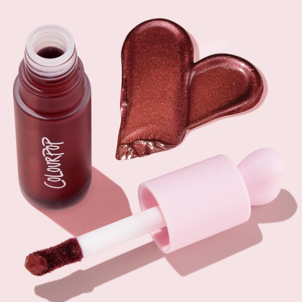 ColourPop Liquid Blush in Wine n' Dine, open frosted burgundy bottle with pink doe-foot applicator swiping a metallic wine-rose heart swatch on a pale pink backdrop, showcasing the shimmery, blendable liquid formula.