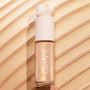 ColourPop Liquid Highlighter in Sugar Donut stands upright with a heart-shaped cap, showcasing a luminous champagne‑gold glow in a frosted tube, set against sweeping metallic swatches that emphasize its shimmery finish.