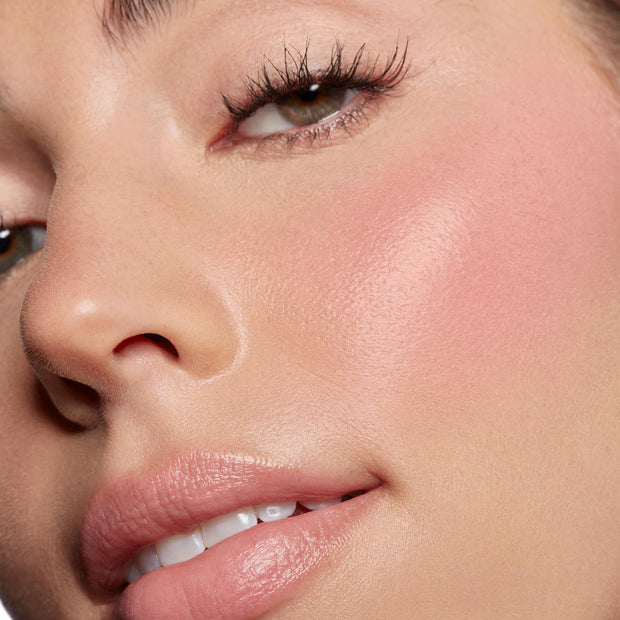 ColourPop Lip and Cheek Balm in Lucky U delivering a dewy soft-pink flush on the cheek and a glossy nude tint on the lips, shown in an extreme close-up of a face with smooth skin and long lashes under natural light.