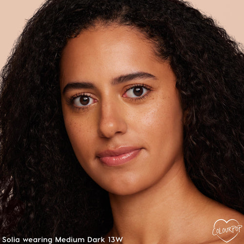 Model wearing ColourPop Pretty Fresh Tinted Moisturizer in Medium Dark 13W (warm), showcasing a natural, dewy, even complexion with light coverage and freckles visible, close-up against a soft beige studio background.