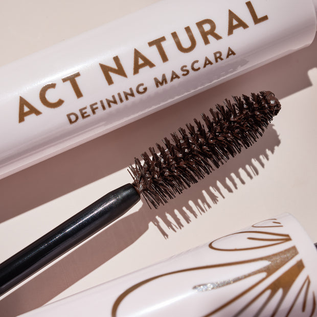 Mascara wand coated in rich brown formula rests beside a soft pink tube reading Act Natural Defining Mascara, highlighting dense spoolie bristles; ColourPop Act Natural Mascara in Brown shown on a neutral backdrop.