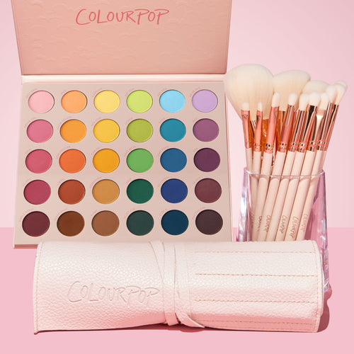 Crazy For Hue ColourPop Shadow Palette & Brush Set displays a 30-pan matte rainbow palette with an open pink lid, paired with soft white-bristle brushes with rose-gold ferrules in a clear cup and a pale pink embossed brush roll, styled on a pink backdrop.