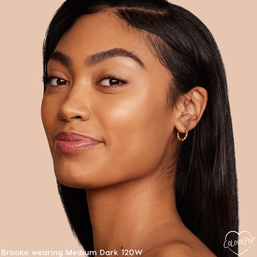 Model showcasing Medium Dark 120W shade, wearing ColourPop Pretty Fresh Creamy Concealer for a smooth, radiant natural finish; close-up side profile with glossy lips and small gold hoop earring against a soft beige background.