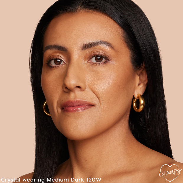 Model wearing ColourPop Hyaluronic Hydrating Foundation in Medium Dark 120W, showing even, natural coverage with a soft glow; close-up face with gold hoop earrings against a warm beige background.