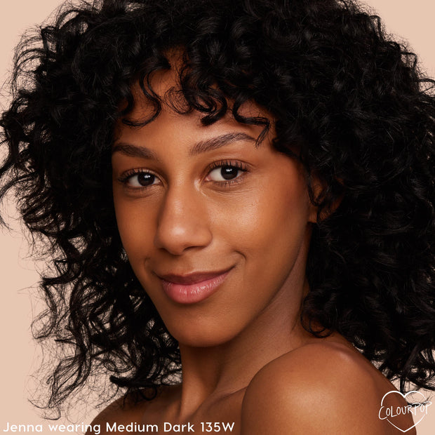 Model wears ColourPop Hyaluronic Hydrating Foundation, shade Medium Dark 135W, showcasing hydrated, dewy medium coverage with warm undertones; tight headshot against a neutral beige background for shade match reference.