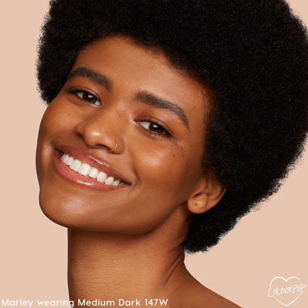 Model smiling in close-up, wearing ColourPop Hyaluronic Hydrating Foundation in Medium Dark 147W, showcasing dewy, even coverage against a warm beige backdrop to highlight the shade match and skin-like finish.