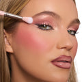 Model eye wearing rosy shimmer from the Smoke 'N Roses ColourPop Shadow Palette, makeup brush blending pink metallic shadow at the outer lid, paired with soft blush and a mauve lip, studio close-up on white background.