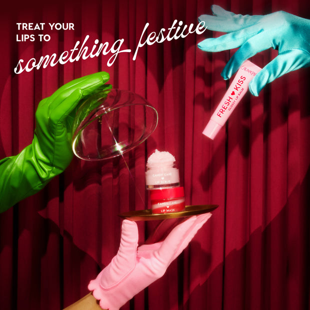 Mistletoe Kisses lip care kit by Colourpop - Fresh Kiss glossy lip balm and Candy Cane Lip Scrub + Lip Mask - presented by green, blue, and pink gloved hands lifting a glass cloche over a gold tray against a red holiday curtain.