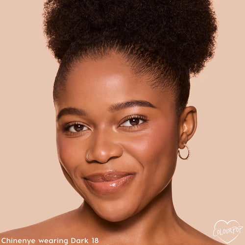 Model wearing ColourPop Pretty Fresh Tinted Foundation Balm in Dark 18, smiling in a studio close-up that showcases smooth, even, dewy skin with natural coverage, small hoop earring visible, against a beige backdrop for accurate shade reference.