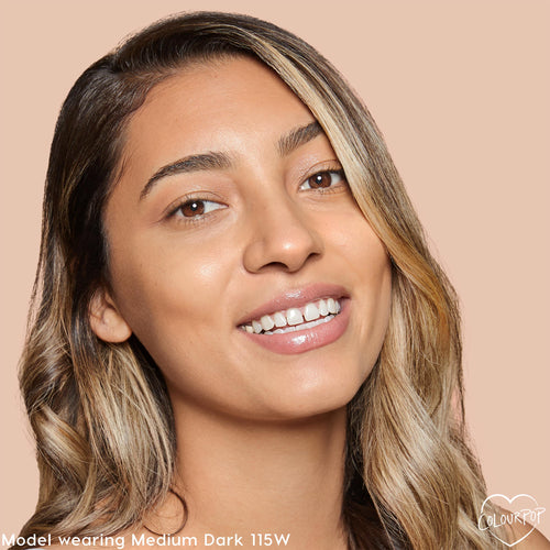 Model smiling wears ColourPop Pretty Fresh Creamy Concealer, shade Medium Dark 115 W (warm), showcasing smooth medium-dark coverage and a dewy finish on even-toned skin against a soft beige background.